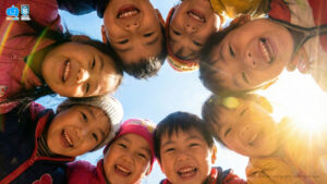 A low-angle shot of a group of joyful kindergarten children looking down into the camera in a circle under a bright, sunny sky.